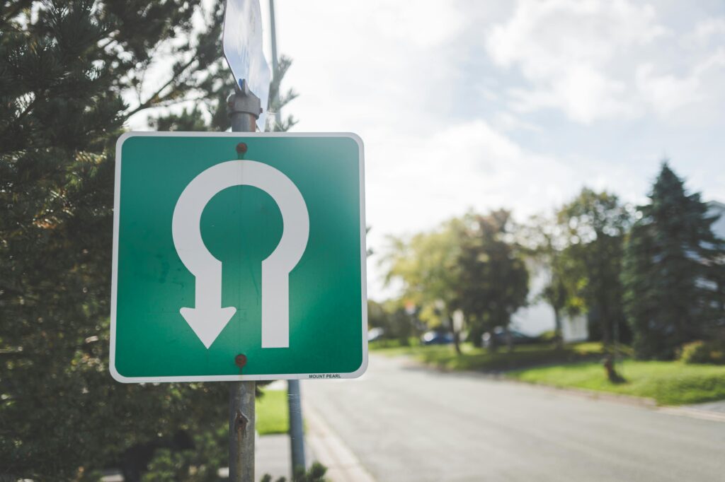 Close-up of a green U-turn road sign on a sunny suburban street with trees.
